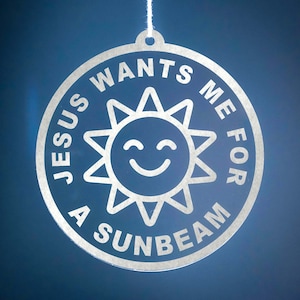 May include: Circular silver-colored ornament with the text "JESUS WANTS ME FOR A SUNBEAM" encircling a smiling sun design. The ornament is suspended by a white string against a blue background. Ideal for religious-themed decor.
