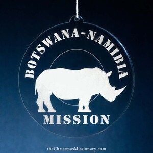 May include: Clear, circular ornament with the text "BOTSWANA-NAMIBIA MISSION" and a rhino silhouette. The ornament is suspended by a white string against a dark blue background. The website address "theChristmasMissionary.com" is at the bottom.