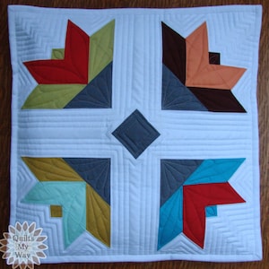 May include: A white pillow with a geometric design featuring four large, multi-colored stars. The stars are made up of red, orange, teal, green, brown, and gray fabric. The pillow is quilted with a diamond pattern in the center and straight lines around the edges.