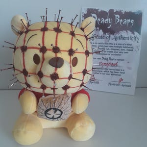 May include: A plush yellow bear with a red grid pattern and numerous metal nails inserted into its head. The bear is holding a small, tattered bag. A certificate of authenticity is visible in the background.