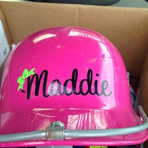 Puede incluir: Casco de b&eacute;isbol rosa con una pegatina negra y verde que dice "Maddie".