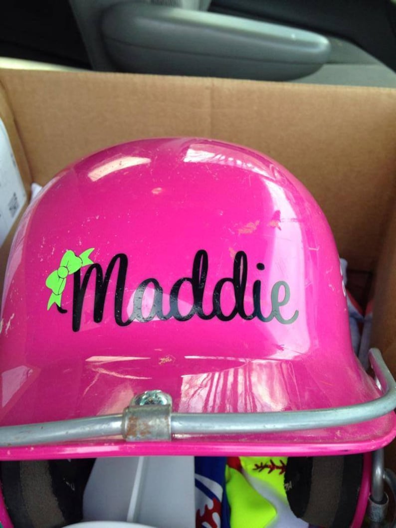 Puede incluir: Casco de b&eacute;isbol rosa con una pegatina negra y verde que dice "Maddie".