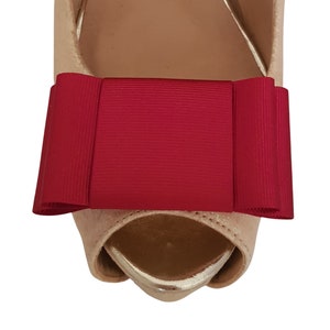 May include: A close-up of a pair of beige high-heeled shoes with a large red bow on the front.