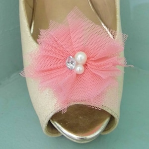 Puede incluir: Primer plano de un zapato peep-toe color crema adornado con una flor de tul rosa. La flor está adornada con un brillante transparente y dos perlas blancas. El zapato tiene un ribete metálico dorado.
