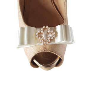 Könnte beinhalten: Ein Paar beige Peeptoe-Heels mit einer weißen Schleife aus Satin und einem perlverzierten Kreuz als Schmuckstück.