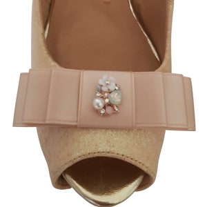 Puede incluir: Un par de tacones peep-toe de brillo dorado claro con un lazo de satén rosa y un adorno de piedras preciosas.