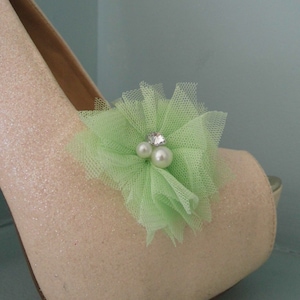Puede incluir: Un par de tacones altos de brillo rosa claro con un adorno de flor de tul verde. La flor tiene un pequeño strass y detalles de perlas.