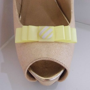 Pode incluir: Um par de saltos de bico aberto bege claro com um laço amarelo e um botão listrado branco e amarelo.