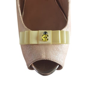 Pode incluir: Um par de saltos peep-toe rosa claro com um laço de cetim amarelo e um enfeite de joaninha.