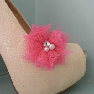 Puede incluir: Un par de tacones altos de brillo color crema con un adorno de flor de tul rosa. La flor tiene un centro de pedrería y perlas.