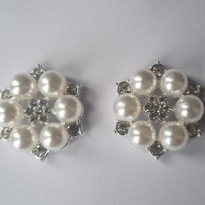 May include: A pair of silver-toned, floral-shaped earrings. Each earring features six large, round, white faux pearls surrounding a central cluster of smaller, clear crystals. Additional clear crystals are set around the outer edges.