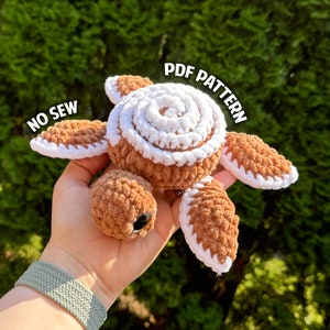 Puede incluir: Una tortuga de crochet marrón y blanca con una concha blanca y extremidades marrones. La tortuga está siendo sostenida en una mano. El texto "NO SEW" y "PDF PATTERN" son visibles en la imagen.