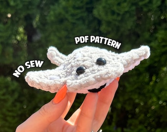 May include: A crocheted gray manta ray with black eyes and a white mouth. The manta ray is being held by a hand with orange nail polish. The text "PDF PATTERN" and "NO SEW" are visible in the image.