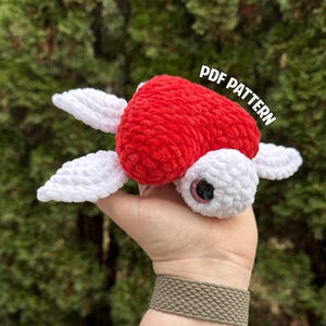May include: A crocheted red and white turtle amigurumi toy with a large eye. The toy is being held in a hand with a brown bracelet. The text "PDF PATTERN" is visible in the background.