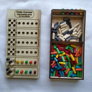 May include: A wooden game board with the title "Code Cracker" and the text "Code Breaking game for two players". The board has six rows of holes and a row of pegs at the bottom. The game includes a wooden box with three compartments filled with colourful wooden pegs.