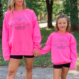 May include: Two pink sweatshirts with black text and hand drawn hearts. The larger sweatshirt says "Ellie Dance Big Sister Audrey" and the smaller sweatshirt says "Ellie Dance Little Sister Audrey".