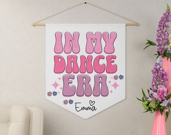 May include: A white banner with pink text that reads "In My Dance Era" with a heart and the name "Emma" below. The banner is hanging from a wooden dowel rod.