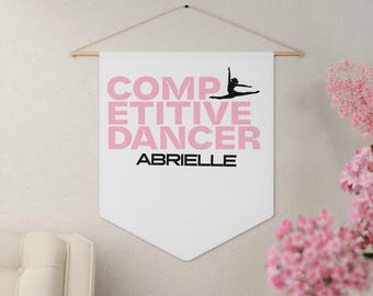 May include: A white banner with the words "COMPETITIVE DANCER" in pink block letters and the name "ABRIELLE" in black. A black silhouette of a dancer is above the word "ABRIELLE". The banner is hanging from a wooden rod.