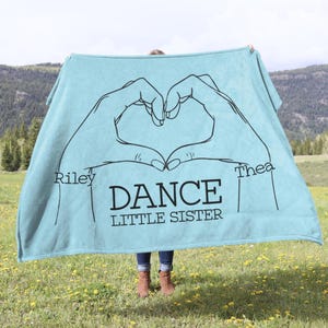 May include: A light blue fleece blanket with a heart shape formed by two hands. The text "DANCE LITTLE SISTER" is printed in black on the blanket. The names "Riley" and "Thea" are printed on either side of the heart.