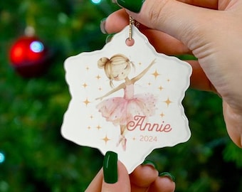 May include: A snowflake-shaped Christmas ornament with a ballerina in a pink tutu. The ornament is printed with the name "Annie" and the year "2024". The background is a blurred Christmas tree.