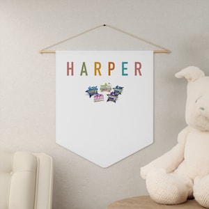 Puede incluir: Una pancarta blanca con el nombre "HARPER" en letras de arcoíris cuelga de una varilla dorada. Cinco pines de estrella de colores con palabras relacionadas con la danza están unidos a la pancarta.