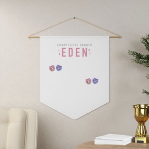 Peut inclure: Bannière blanche avec le texte "COMPETITIVE DANCER *EDEN*" en lettres roses. Deux petites épingles colorées sont attachées à la bannière.