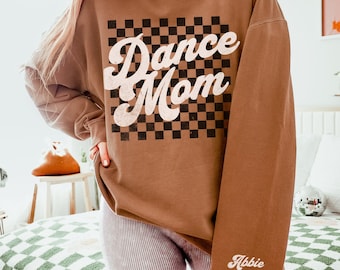 May include: A brown sweatshirt with a checkered pattern and the text "Dance Mom" in a retro font. The sweatshirt has long sleeves and a crew neck. The sleeve has the name "Abbie" written on it.