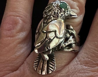 Könnte beinhalten: Ein silberner Vogelring mit einem smaragdgrünen Steinauge. Der Ring ist mit strukturierten Federn und einem astähnlichen Band versehen. Der Vogel sitzt auf dem Finger und zeigt das aufwendige Design und die Handwerkskunst des Schmucks.