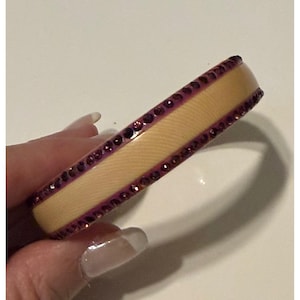 May include: A bangle bracelet featuring a smooth, light beige center band flanked by two rows of small, dark purple circular stones. The bracelet has a rounded profile and a consistent width throughout its circular structure.