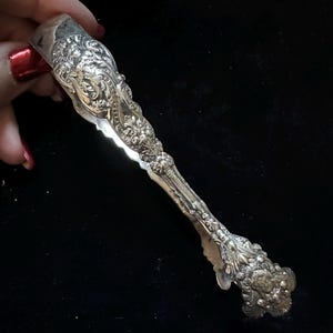 Puede incluir: Pinzas de azúcar plateadas y ornamentadas con diseños florales y de volutas detallados. Las pinzas están sujetas por una mano con esmalte de uñas rojo. Es un artículo vintage.