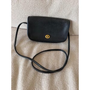 May include: A black leather crossbody bag with a rectangular shape and rounded corners. The bag features a gold-tone metal turn-lock closure on the front flap and a thin, matching black leather shoulder strap attached to the sides of the bag.