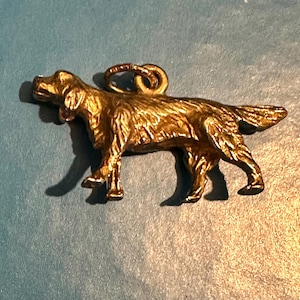 May include: A gold-coloured pendant in the shape of a dog, possibly a retriever, with a detailed textured surface. The dog is standing with its tail up and a loop at the top for a chain.