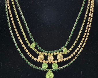 May include: A multi-strand necklace with green and gold rhinestones. The necklace has a graduated design with larger rhinestones in the center and smaller rhinestones on the sides. The necklace is finished with a single, large, green rhinestone pendant.