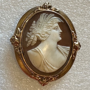 Puede incluir: Un broche de camafeo dorado con una talla de camafeo blanco de un perfil de mujer con un tocado de plumas. El camafeo está engastado en un marco ovalado con detalles florales.