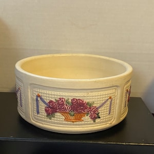 Puede incluir: Un tazón de cerámica blanco con un diseño floral. El tazón tiene una cesta de rosas rosadas con cintas moradas y un fondo blanco.