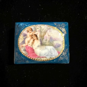May include: A rectangular, blue trinket box with a painted oval scene of cherubs and a woman in a flowing gown. The oval is framed with gold trim and the box has gold filigree accents.
