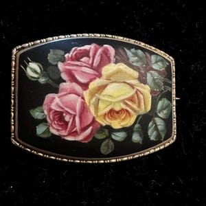 May include: A rectangular brooch with a painted floral design. The brooch features pink and yellow roses with green leaves against a black background, framed by a gold-colored metal border.