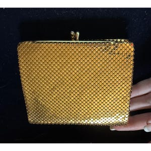 May include: A rectangular gold-toned metal mesh clutch purse featuring a fine, interlocking chainmail texture. The bag is secured at the top with a metallic kiss-lock closure consisting of two small, rounded knobs that snap together firmly.