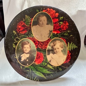 May include: A round, black, decorative plate with three oval portraits of children. The portraits are surrounded by red roses and green ferns. The plate has a gold rim and the text "D. Siepfer" in the bottom right corner.