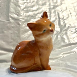 以下が含まれることがあります： 座っている猫の陶器製置物。猫は光沢のある温かみのある茶色で、お腹は明るく、小さく尖った耳をしています。青い目とピンクの鼻をしています。装飾品です。