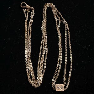Könnte beinhalten: Eine Vintage-Uhrkette in Goldtönen mit mehreren Strängen. Die Kette hat an einem Ende einen Verschluss und am anderen ein rechteckiges Dekorationselement mit kleinen roten und weißen Akzenten. Die Kette wird vor einem schwarzen Hintergrund gezeigt.