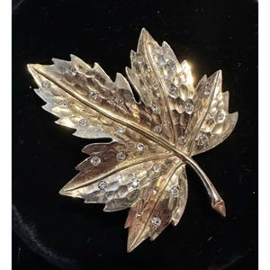 Op de afbeelding: Goudkleurige blad-vormige broche met een getextureerd oppervlak en ingebedde heldere kristallen. De broche heeft een gedetailleerd ontwerp, met de nerven van het blad zichtbaar. De kristallen zijn verspreid over het blad.