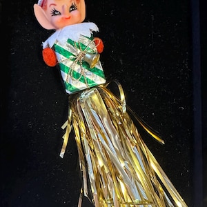 Puede incluir: Adorno navideño vintage con un elfo con gorro rojo y un regalo a rayas verdes y blancas. El adorno tiene una campana dorada y oropel dorado. La cara del elfo está pintada con mejillas rosadas y pestañas oscuras.
