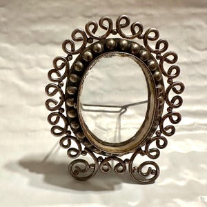 Op de afbeelding: Een antiek zilverkleurig ovaal fotolijstje met een decoratief, krullend ontwerp. Het frame heeft een helder glazen inzetstuk en een kleine standaard. Het ontwerp van het frame bevat kleine, ronde kralen rond de ovale opening.