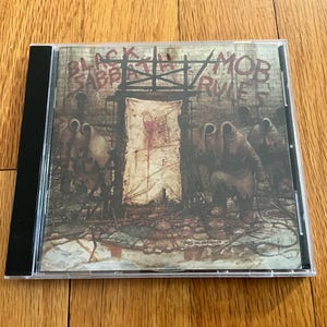 May include: A CD case featuring the album cover for Black Sabbath's "Mob Rules." The artwork depicts a dark, ominous scene with figures in hooded cloaks and the album title in a distressed font.