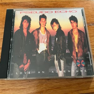 May include: CD case for the album "Love An Adventure" by Pseudo Echo. The cover features four men in leather jackets and casual attire against a sunset backdrop. The album title and band name are printed in red.