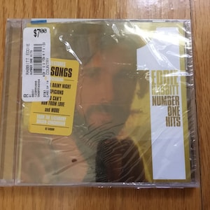 May include: A gold CD case featuring a picture of Eddie Rabbitt on the front. The case is wrapped in plastic and has a price tag on the top left corner. The title of the CD is "Eddie Rabbitt Number One Hits".