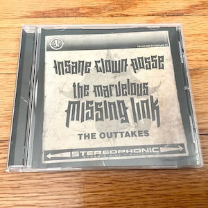 May include: A CD case featuring the album "The Marvelous Missing Link: The Outtakes" by Insane Clown Posse. The album art has a distressed, vintage look with black text on a beige background. The case is clear plastic.