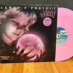 May include: A pink vinyl record with a black and white album cover featuring a young boy holding a glowing pink globe. The album cover has the title "Planet P. Project Pink World" and the text "Behind the Barrier - What I See - Power".