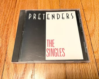 May include: A CD case for the Pretenders' album "The Singles." The album cover features the band's name in white, stylized font above the title in pink. The case is black and clear.